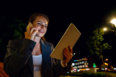 Businesswoman multitasking with phone and tablet during nighttime walk in cityの写真素材