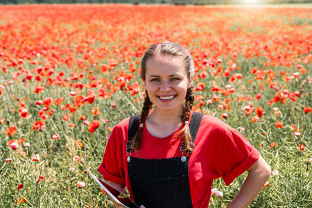Joyful young woman ecological farmer with tablet enjoying summer day in blooming red poppy flower fieldの写真素材