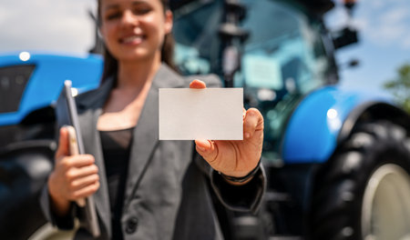 Mockup of blank business card in hand of female agricultural machinery dealer with tractor in backgroundの写真素材