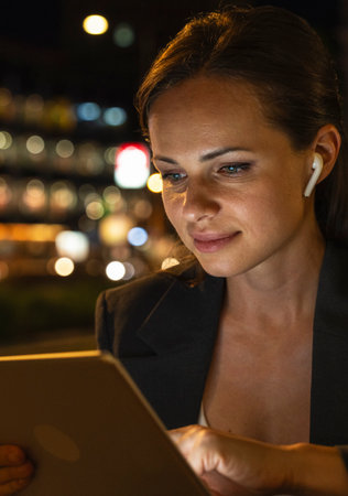 Portrait of modern female entrepreneur with wireless earbuds using digital tablet in night city