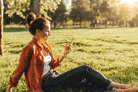 Young woman using smartphone while relaxing on green grass outdoorsの写真素材