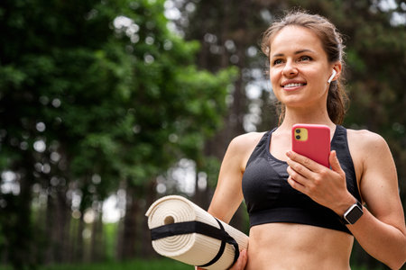 Portrait of modern fit woman with rolled up yoga mat and smartphone in her hands outdoorsの写真素材