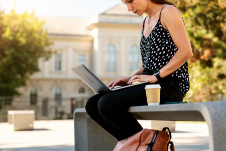 Female using laptop while sitting on bench on city street. Mockup of brown paper coffee cup with copy space.の写真素材