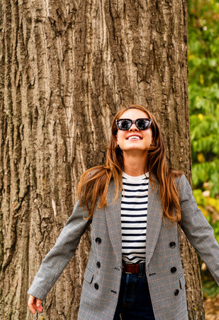 Cheerful brunette female wearing sunglasses posing near the big tree in city park.の写真素材