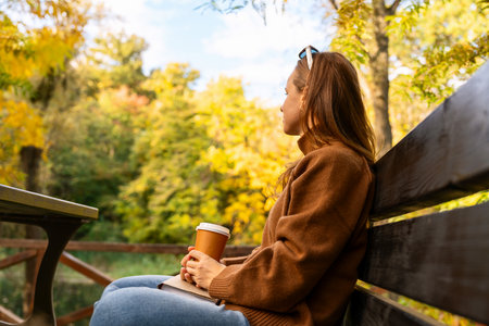 Woman with hot drink in paper cup relaxing in autumn park.の写真素材