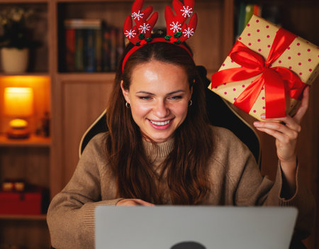 Festive woman in reindeer antlers holding Christmas gift during online family callの写真素材