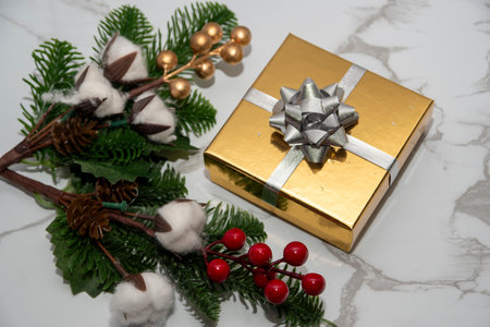 Golden Christmas gift box with silver bow placed beside fir branches, pine cones and red berries on white marble background.の写真素材