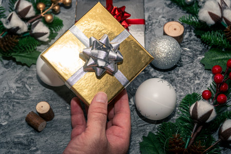Hand offering a golden Christmas gift box with silver bow among baubles and greenery, holiday giving and celebration concept.の写真素材