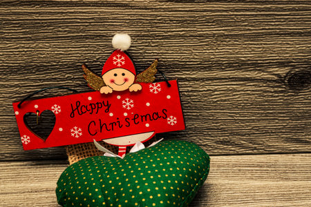 Cute wooden angel holding a red Happy Christmas sign over green cushion on rustic wood, playful holiday greeting decoration.の写真素材