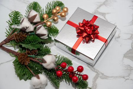 Elegant silver Christmas gift box with red bow next to green branches, berries and cotton on white marble background.の写真素材
