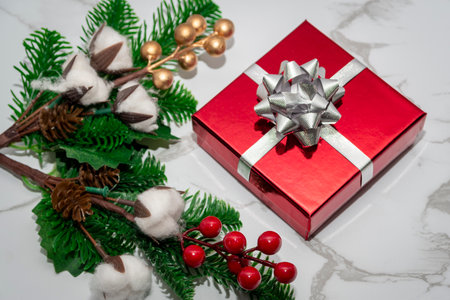 Red Christmas gift box with silver bow surrounded by evergreen branches, cotton and red berries on light marble background.の写真素材