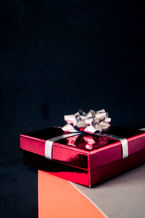 Red present box wrapped with silver ribbon on orange and gray geometric surface, dark background and copy space for holiday, sale or celebration banners.の写真素材