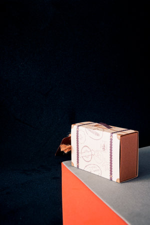 Mini suitcase-style cardboard gift box on orange and gray geometric surface against black background, studio still life with copy space.の写真素材