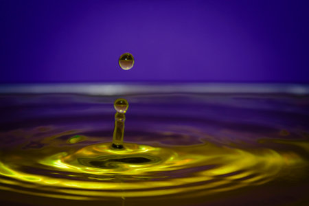 Vibrant yellow water splash with suspended droplet on purple background, creating dynamic abstract liquid art with bright reflections.の写真素材