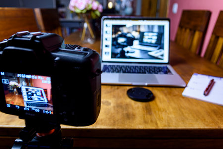 Behind-the-scenes home studio: camera on tripod filming a laptop with photo editing on a wooden table. Cozy creator workspace with notebook.の写真素材