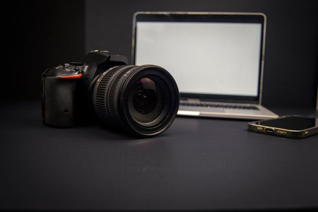 Dark creative workspace with DSLR camera, laptop and smartphone. Blank screens and copy space for...