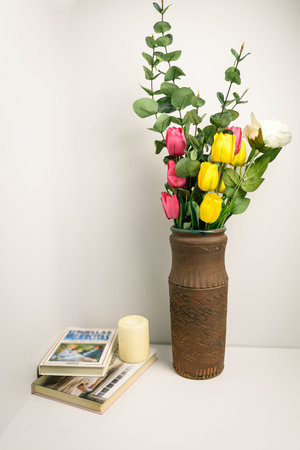 Colorful tulips and eucalyptus in a rustic terracotta vase with books and candle on a white shelf,...