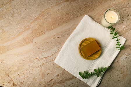 Top view of white towel with soap on wooden dish and lit candle on beige marble, decorated with...