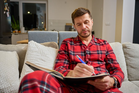 Man sitting on sofa in pajamas and writing in notebook at homeの写真素材