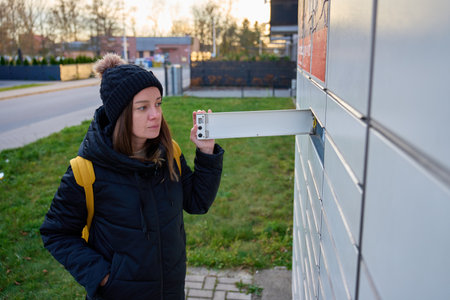 Woman wearing winter jacket and hat opening metal mailbox outside apartment building. Concept of...