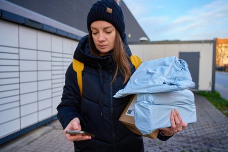 Woman holding multiple parcels and using smartphone near automated parcel lockers. Concept of online shopping and parcel pickupの写真素材