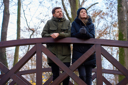 Man and woman leaning on bridge railing in park with trees in autumn. Concept of couple leisureの写真素材