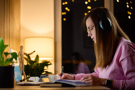 Woman wearing headphones writes in notebook while sitting at desk with laptop in home interior....