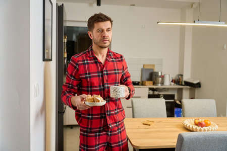 Man in red pajamas standing in kitchen holding mug of coffee and plate with pastries. Concept of evening routineの写真素材