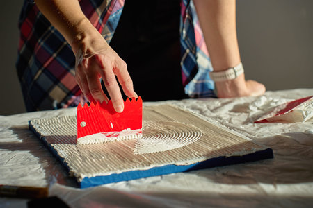 Hand pressing red plastic scraper into thick paste to form curved textured lines on flat board....