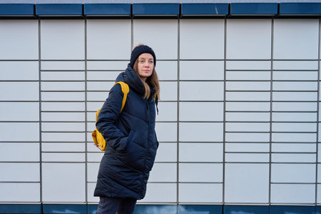 Woman in winter jacket and knit hat standing against wall with backpack. Concept of urban lifestyle