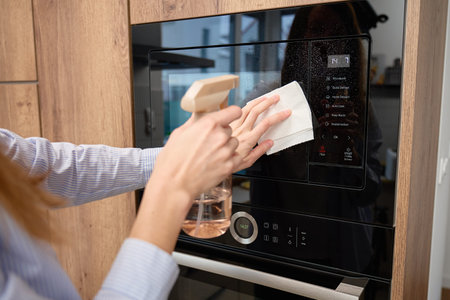 Hands spray and wipe glass door of built in microwave oven in kitchen cabinet
