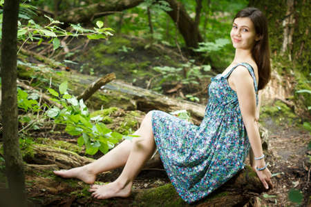Beautiful young woman wearing elegant white dress walking on a forest path with rays of sunlight beaming through the leaves of the treesの写真素材