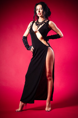 Beautiful woman in black dress and stockings posing at camera against red backgroundの写真素材