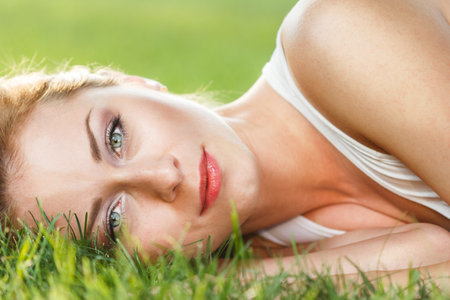 Close up portrait of an attractive young woman laying down on green grass in a park during the summer and listening to music with her earphones, being thoughtful outdoors.の写真素材