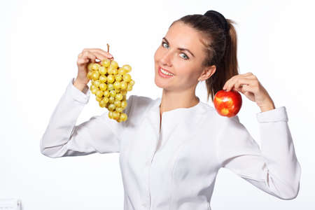 young attractive woman, holding a grape in one hand and a red apple in the other, looking joyful and happy, standing over isolated white background.の写真素材