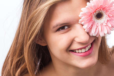 Portrait of a happy girl with fresh skin holding a flower over one eye on white background. Close up portrait of beautiful redhead curly woman pnik flower wearing white tshirt. Beauty and springの写真素材