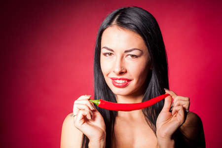Beautiful woman in black lace lingerie posing in studio with red background. She is playing with chili pepper.の写真素材