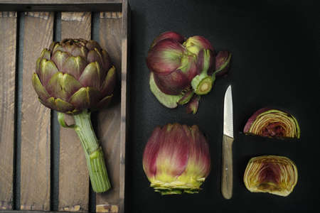 Fresh artichoke green-purple flower head on cutting board with knife on black backgroundの写真素材