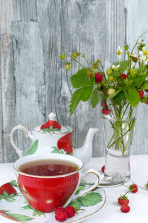 Fresh wild strawberry tea in an colorful cup and teapot and bouquet of ripe wild strawberries  in a vase on wooden backgroundの写真素材