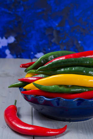 Red, green and yellow mexican hot chili peppers on blue background in blue bowl
