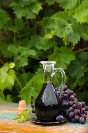 Black old balsamic vinegar in a glass jug with fresh red grapes on green vineyard background on wooden table, outsideの写真素材