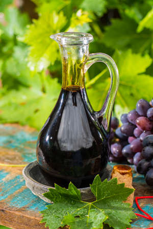 Black old balsamic vinegar in a glass jug with fresh red grapes on green vineyard background on wooden table, outsideの写真素材