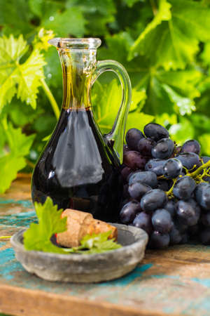 Black old balsamic vinegar in a glass jug with fresh red grapes on green vineyard background on wooden table, outsideの写真素材