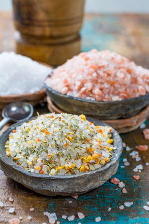 Different types of natural salt in stone bowls on wooden surface. White sea salt, pink Himalayan salt, Spiced salt with rosemaryの写真素材