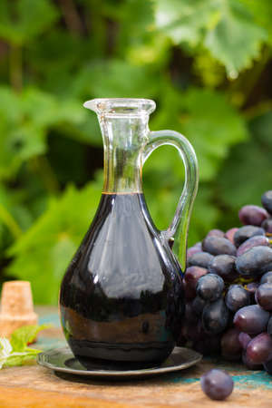 Black old balsamic vinegar in a glass jug with fresh red grapes on green vineyard on wooden table, outsideの写真素材
