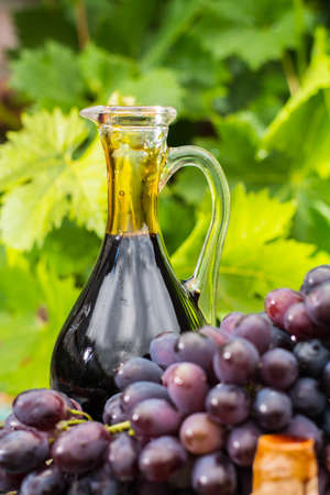 Black old balsamic vinegar in a glass jug with fresh red grapes on green vineyard background on wooden table, outsideの写真素材