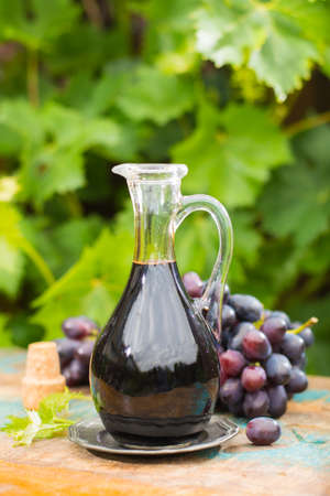 Black old balsamic vinegar in a glass jug with fresh red grapes on green vineyard background on wooden table, outsideの写真素材