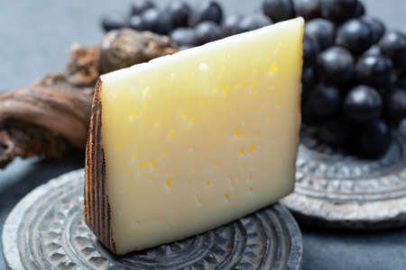 Piece of Spanish hard scheep milk cheese Manchego served with black grapes close up