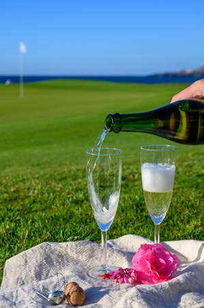 Pouring of bubbles white champagne or cava wine in two glasses during romantic event or celebration on green golf club grass with sea viewの写真素材