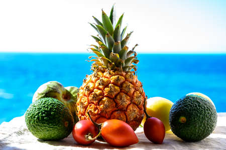 Tropical fruits collection, custard apple or green ripe cherymoia, passion fruit, pineapple, mango, tamarillo, avocado exotic fruits on blue sea background in sunny dayの写真素材